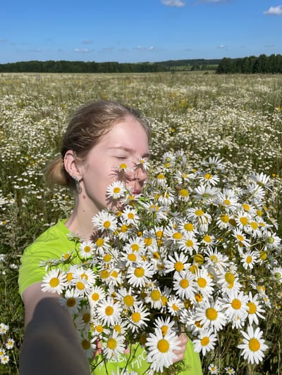 A young woman with her eyes closed is holding a large bouquet of white daisies in a field of similar flowers. She is wearing a bright green t-shirt. The background shows a vast field of daisies under a blue sky with scattered clouds, and a distant line of trees. The location is identified as Сям-Можга, Russia.