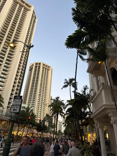 The image, rotated counterclockwise, depicts a street scene in Honolulu, USA. Two tall buildings with repetitive architectural designs dominate the view, framed by palm trees and a blue sky. A street sign indicates a bike lane ahead and a one-way street. A crowd of people is visible on the right side of the image.