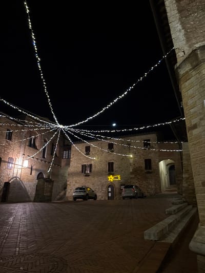 A nighttime view captures a building decorated with strings of lights radiating outward, forming a festive display. The building features brickwork and several windows, with a lit star hanging on one wall. Two cars are parked nearby. The sky is a dark contrast to the brightness of the lights and a faint crescent moon can be seen. The shot is taken from a low angle, looking upwards between the building and an archway.