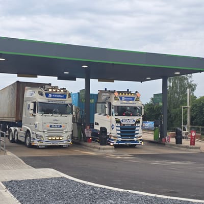 This image shows two large trucks parked under a petrol station canopy. The truck on the left is a white DAF, and the truck on the right is a white and blue Scania. Both trucks are carrying large shipping containers. The petrol station has several pumps visible, and a sign for Greggs bakery is also present. The sky is overcast, and trees are visible in the background. The location appears to be a services area or truck stop. There are no people visible in the immediate vicinity of the trucks. The presence of the Greggs sign suggests a location in a town or city where such amenities are common, and the mention of "Huntingdonshire, United Kingdom" in the prompt implies this specific location.
