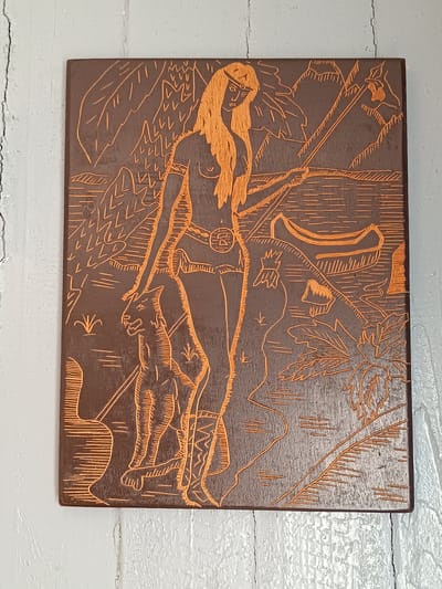 The image shows a carved wooden plaque depicting a scene with a woman and a feline creature. The woman has long, flowing hair and wears a headband. She is partially undressed, with her breasts exposed. A belt with a circular emblem cinches her waist. She stands next to a large, quadrupedal animal, possibly a panther or a jaguar, which has a stylized appearance. The background suggests a natural landscape with mountains, water, and palm trees. A canoe is visible on the water. The carving is done in relief, with the design etched into the wood, creating an orange-brown contrast against the darker wood. The plaque is mounted on a white, textured wall with visible cracks. The image does not provide any information about the location of Tebras, Latvia.