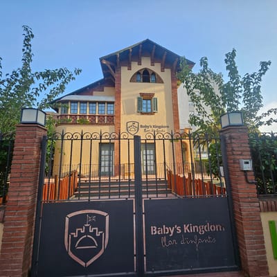 This media file features a frontal view of a multi-story building with a distinctive architectural style, possibly dating from the early 20th century, located in Girona, Spain. The building has a light yellow facade, a dark tiled roof with decorative eaves, and a prominent arched window on the upper level with a black metal grate. The ground floor features two sets of double doors with windows. The building is fronted by a black metal gate with decorative scrollwork. The gate displays the name "Baby's Kingdom" in large, stylized text, accompanied by the Catalan phrase "llar d'infants" below it, indicating it is a nursery or kindergarten. A crest featuring a castle-like structure with a flag is also visible on the left side of the gate.
The scene is set during daylight, likely in the late afternoon or early evening, as suggested by the warm lighting and long shadows. A sliver of the moon is visible in the clear blue sky, indicating it's not quite dusk. Lush green trees flank the building and parts of the gate, suggesting a well-maintained property. The overall impression is one of a charming and established educational institution. There are no people visible in the image. The weather appears clear and calm.