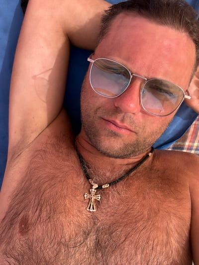The image depicts a scene in Bhagatwadi, India. An adult male is shown in a close-up, high-angle shot, lying in a reclined position, appearing to rest outdoors. He has short dark hair, visible stubble on his face, and prominent chest hair. His skin displays a tanned and slightly reddened complexion. He wears silver-framed aviator-style sunglasses with light, reflective lenses. Around his neck is a dark, braided necklace adorned with a detailed gold-colored cross pendant. His right arm is bent with the hand positioned behind his head or neck. The background indicates he is resting on a surface covered by blue fabric on the left and a red, white, and blue/green plaid fabric on the right.