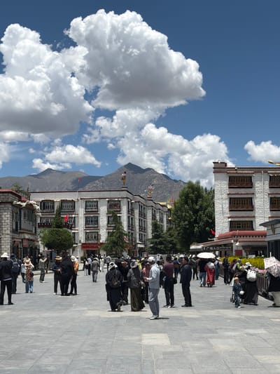 This image captures a vibrant public square or street scene in Chengguan District, China, under a clear, bright blue sky with large, fluffy white cumulus clouds. The strong sunlight and visible shadows suggest it is midday or early afternoon.
The scene is populated by numerous people walking, standing, and gathering across a wide, paved stone square. Individuals are dressed in a variety of casual clothing, with some carrying umbrellas, likely for sun protection. Their activities suggest everyday movement and congregation in a public space.
To the right of the square, a row of multi-story buildings showcases traditional Tibetan architectural elements, characterized by white-textured walls and dark-framed windows with intricate designs. Green trees are strategically placed near these structures, adding to the landscape. Colorful prayer flags are visible, draped from buildings and on the distant mountainside, serving as a prominent cultural cue.
In the background, a range of majestic, rugged mountains extends under the expansive sky. A red flag is seen flying from one of the buildings. A significant textual detail is a sign on a building that reads "广场入口" (Guǎngchǎng rùkǒu), which translates to "Square Entrance." The overall impression is one of a busy yet serene environment, reflecting the daily life and cultural richness of the area.