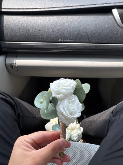 In Imeni Chapaeva, Kyrgyzstan, a person is holding a bouquet of artificial white roses and eucalyptus stems. The bouquet is tied with twine. The person is wearing black pants. The background appears to be the interior of a car, with the dashboard and a glove compartment visible. The lighting suggests it is daytime.
