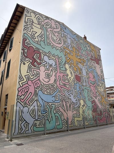 This is a media file depicting a vibrant mural on the side of a building. The mural features numerous stylized figures, characteristic of Keith Haring's art, rendered in pastel colors such as yellow, pink, light blue, light green, and burgundy, all outlined in black. The figures are depicted in dynamic, contorted poses, appearing to dance, leap, and interact with each other, creating a sense of movement and energy. Some figures are abstract and fluid, while others have more defined limbs and heads.

In the center of the mural, there is a distinct object: a red rectangular shape with three horizontal lines and a circular button on it, resembling a control panel or a stylized television. Surrounding the figures are numerous black, wavy lines, adding to the overall chaotic yet harmonious composition.

The mural covers a large portion of the building's exterior. The building itself appears to be multi-story, with some windows visible on the lower section. The building's facade below the mural is a pale yellow, with wooden beams and dark grates for ventilation. The mural is oriented vertically on the side of the building, and the photograph is taken from a low angle, looking upwards.

To the left of the mural, the sky is visible, with the sun shining brightly, creating a lens flare. The weather appears to be clear and sunny, suggesting it is daytime. A paved area, possibly a street or sidewalk, is visible to the right of the building, bordered by a gray textured wall.

The context provided indicates the location is Pisa, Italy. The overall emotion conveyed by the artwork is one of joy, exuberance, and connection through movement. There is no visible text in the mural itself, aside from the stylized red object which could be interpreted as symbolic.
