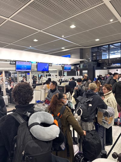 An airport departure area is packed with passengers and their luggage. Individuals stand or sit in line, waiting. Several people are focused on their phones. Large digital displays on the wall indicate the status of flights at gates B22, B23, and B24, indicating "Economy Class". The scene suggests a crowded travel hub with people anticipating their upcoming flights. The camera angle is slightly tilted.