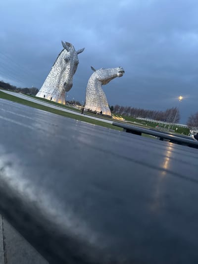 The scene depicts The Kelpies, two colossal equine head sculptures, located in The Helix Park, Falkirk, United Kingdom, at dusk or early evening. The two metallic sculptures, constructed from numerous steel plates, rise prominently from a landscaped green area. The sculpture on the left faces partially towards the viewer, exhibiting a more upright neck, while the one on the right faces right, with its neck in a more downward, arching posture. Both sculptures are illuminated from their bases with warm light.
The sky above is overcast and dark grey-blue, indicative of twilight. The ground consists of manicured grassy lawns and paved pathways. Several small, indistinct human figures are visible near the bases of both sculptures, appearing to be observing or walking around them. One figure stands near the left sculpture's base, and multiple figures are discernible near the right sculpture's base.
In the mid-ground, a line of slender, bare-branched deciduous trees is visible, extending towards the right. A tall street light with an illuminated fixture stands further right. The immediate foreground is dominated by a dark, flat, and horizontally textured surface, likely the top of an outdoor table or bench, which occupies approximately the lower half of the image. Reflections of ambient light are visible on this surface. The perspective is a low-angle shot looking up towards the sculptures.