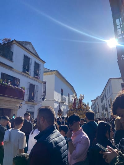 A street-level view captures a religious procession in Córdoba, Spain. Numerous spectators line the street, observing the passing float. The float carries religious figures adorned with white garments. Buildings with white facades and wrought-iron balconies rise on either side of the street. The sky is blue and the sun is shining brightly.