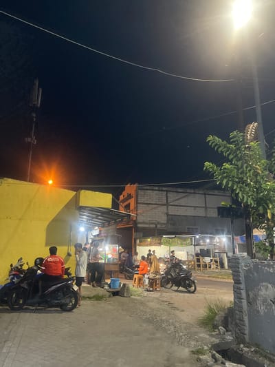 The image depicts an outdoor scene at night, likely in Anggaswangi, Indonesia, where a group of people are gathered around food stalls. Several motorcycles are parked on a paved area in the foreground. A bright street lamp illuminates the scene from the upper right. In the background, there are low-rise buildings, some of which appear to be shops or eateries, with lights on.
Specifically, there are three motorcycles on the left side of the image. A man wearing a red jacket with "Shopee Food" written on the back is seated on one of the motorcycles. To his right, two men are standing. Further to the right, near the food stalls, several individuals are visible. One woman is wearing an orange headscarf and seated on a stool, with another person next to her. Other figures are seen standing or moving around the food stalls.
The environment includes a yellow wall on the left, a paved walkway, and a road in the middle ground. The sky is dark, indicating it is nighttime. A cell tower is visible on the left side of the frame. The overall atmosphere suggests a lively, communal gathering at a night market or street food area.