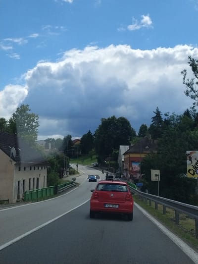 The image shows a rural road in Dražice, Czechia, on a cloudy day. The road is paved and has a white line marking the lane. On the left side of the road, there is a light-colored building with a dark roof and a satellite dish. There is also a green metal fence along the side of the road. On the right side of the road, there is a guardrail and trees. Several cars are visible on the road, including a red Volkswagen Polo. In the background, there are houses and trees. The sky is blue with large white clouds. A sign with "Cukrárna" (confectionery in Czech) is visible on the right side.