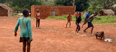 The scene depicts an outdoor residential area in Kundu 1, Democratic Republic of the Congo, characterized by reddish-brown dirt ground and basic structures. Eight children, appearing to be male and ranging in age from toddler to pre-teen, are present.

In the immediate foreground, a child with their back to the viewer wears a light blue, long-sleeved shirt and dark shorts. To the left, a partially obscured hut with a thatched roof is visible. In the mid-ground, a central brick building with an unglazed window opening forms a backdrop. Several children are gathered to the right of this building. One child in a patterned orange and red shirt stands to the left, while another in a similar patterned short-sleeved shirt stands further right. Towards the right side of the frame, three children are in active postures: one in a black long-sleeved shirt with striped pants leans forward, another in a grey tank top and dark shorts is lunging forward, and a child in a blue t-shirt and dark shorts stands behind them. A toddler in a brown patterned outfit is squatting or crawling on the ground, interacting with a small puddle and debris. Another child in a red top is partially visible in the background, observing. The background features include green foliage, additional thatched huts, and distant hills with more dwellings.