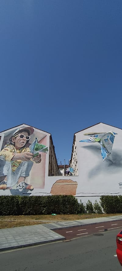 This is a low-angle, vertical photograph of two buildings with murals on their facades in the city of Siero, Spain. The sky is a clear, bright blue.

On the left building, a mural depicts a young child wearing a yellow shirt, light blue overalls, a colorful baseball cap, and white sunglasses. The child is crouching and holding a folded green and white object, possibly a toy or papercraft, with both hands. The mural is rendered in a realistic style with soft shading.

On the right building, another mural shows a three-dimensional illusion of an airplane or kite made of colorful paper or plastic, appearing to fly off the wall. The object is predominantly white, blue, and green, with some text that is partially legible, including "SIER".

The buildings are positioned close together, with a narrow alleyway visible between them, where laundry is hanging on a clothesline. Below the murals, a dense green hedge runs along the base of the buildings. In front of the hedge is a sidewalk with gray paving stones and a red bicycle lane with white lines. A portion of a red car is visible on the right side of the frame, on the street. The ground in front of the hedge also includes patches of dry grass and some green refuse bags. The overall scene suggests a public space adorned with street art.