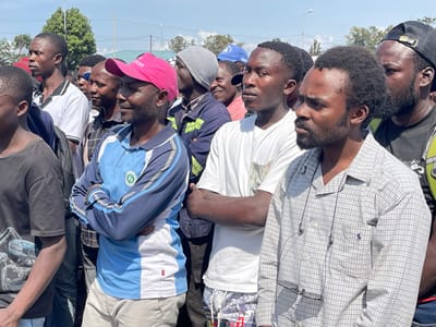 A group of men are standing outside in what appears to be a city setting. The men are all dressed in casual clothing, some have their arms crossed and others are looking straight ahead. The man in the center of the image is wearing a blue and white long-sleeved shirt and a pink hat. The man to the right of him is wearing a white shirt, and the man to the right of him is wearing a white shirt with a floral design. The man to the right of him is wearing a white shirt and black shorts. The man to the right of him is wearing a gray plaid shirt. The man to the right of him is wearing a black shirt. The men are standing in a row, and there is a building in the background. The men appear to be talking, but it's difficult to tell for sure. The location of the photograph is Ville, Democratic Republic of Congo, at latitude -1.663, longitude 29.177.