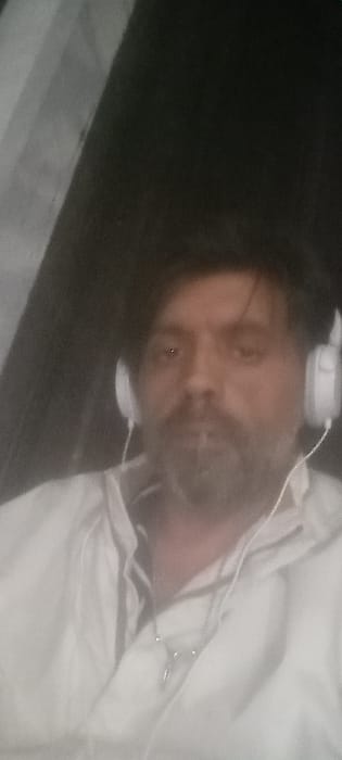 The image displays a close-up, chest-up view of an adult male directly facing the camera. He possesses dark hair and a dark beard with some grey hairs. His eyes appear dark, with a noticeable red-eye effect in the left eye. The individual is wearing white over-ear headphones with visible wires extending downwards. Around his neck, a thin chain or cord with a small pendant is visible. His attire consists of a light-colored, possibly white or off-white, collared garment, which appears to be a shirt or jacket. The background is predominantly dark and out of focus, with a lighter, indistinct vertical element visible on the left side. The scene is dimly lit, suggesting a low-light environment. No identifiable landmarks or features are present within the image to determine a specific location within Montevideo, Uruguay.