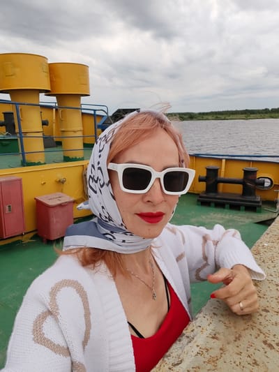 A woman with pink hair, wearing a white patterned scarf, white sunglasses, a white cardigan with beige stripes, and a red top, poses for a selfie on the deck of a ferry. She is leaning on a textured railing. In the background, there are yellow smokestacks, blue railings, and red storage bins on the green deck of the ferry. The ferry appears to be on a body of water, with a tree-lined horizon visible under a cloudy sky. The location is identified as Bereg, Russia.