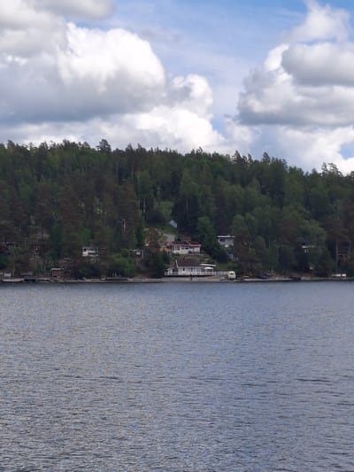 The image displays a waterfront scene situated in Södertälje, Sweden. The lower half of the frame is occupied by a calm body of water, appearing grey-blue with subtle surface ripples. The middle ground features a sloped hillside densely covered with coniferous and some deciduous trees. Approximately ten residential structures are visible on the hillside and along the shoreline. These buildings exhibit varied architectural styles and colors, with several appearing white and others in darker tones. Small piers or docks extend from the shore into the water in front of some properties. A white recreational vehicle, likely a caravan, is parked near the water's edge in front of one house, with a small boat or kayak nearby on the shore. The upper portion of the image reveals a partly cloudy sky, with prominent white cumulus clouds against a light blue background. No individuals or specific actions are discernible.