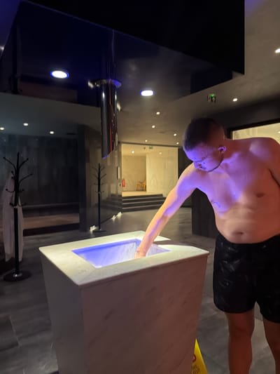 In Hisarya, Bulgaria, a shirtless man with short hair and visible water droplets on his skin is leaning over a white marble basin filled with blue-lit water. He is reaching his left hand into the basin, appearing to test the water temperature. The man is wearing black patterned swim shorts. The environment suggests a spa or wellness facility, with dark tiled flooring, modern lighting including blue accents on the ceiling, and a large reflective metallic cylinder suspended from above. In the background, there are coat racks with white robes, dark walls, and stairs leading to a brighter area with a wooden chair and a fire extinguisher. The lighting casts a purple hue on the man's upper body.