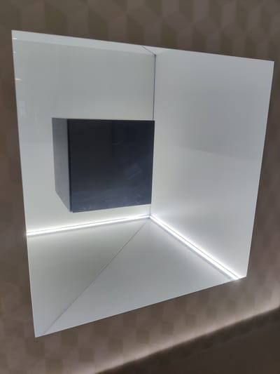 A dark, rectangular object is centrally suspended within a brightly lit, white, geometric display case. The display case has a backdrop that appears to recede into a corner, with angled white planes meeting at a point. LED strip lighting illuminates the edges of the display case, creating a halo effect and casting subtle shadows. The object itself is dark, possibly black or a very dark gray, with a matte finish and some faint scuff marks visible. The surrounding environment appears to be a wall with a textured pattern, possibly wallpaper or a painted finish, in a neutral brown tone. The overall impression is one of modern, minimalist exhibition design. This display is located in the Thessaloniki Museum of Photography, Greece.
