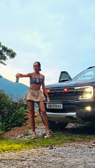 A full-body vertical shot captures a young woman standing on a gravel road beside a black pickup truck, holding a green smartphone outstretched, seemingly taking a selfie or video. She wears a grey strapless crop top, a light brown ruffled mini-skirt, and light pink strappy sandals. Sunglasses are tucked into her top, and she has visible tattoos on her left arm. Her braided hair is styled in several thin strands down her front.
The black pickup truck, identifiable by the "FORD" lettering on its grille and prominent headlights, has its headlights on, emitting a warm glow. Red lights are visible on the grille above the license plate. The Vietnamese license plate reads "29K-179.83". The driver's side door of the truck is open.
The scene is set in a mountainous, rural environment, consistent with Nậm Dịch Commune, Vietnam. The ground is a mix of gravel and dirt, with patches of green grass and bushes on the left and in the mid-ground. Distant green mountains rise in the background under an overcast or twilight sky, suggesting late afternoon or early evening. The soft, diffused light creates a serene outdoor atmosphere, hinting at a stop during a journey or an adventure.