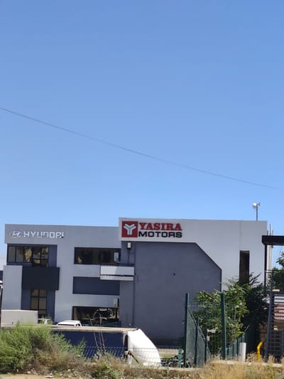 The image shows a modern commercial building with two distinct logos displayed prominently on its facade. On the left side, the "HYUNDAI" logo is visible in silver, featuring the stylized "H" within an oval. The building itself is light gray with dark gray accents and rectangular windows.

To the right of the Hyundai section, a red and black "YASIRA MOTORS" logo is displayed. This section of the building is white with a large, dark gray angled section. A small white antenna or communication device is mounted on the roof of the Yasira Motors section.

In front of the building, there is a lower section with dark windows and what appears to be a white car partially visible. A chain-link fence runs along the front of the property, separating it from a dry, grassy area with some scrubby bushes. The sky is a clear, bright blue, suggesting a sunny day. The overall impression is that of a car dealership or automotive service center.

The location is identified as Atameken, Kazakhstan.