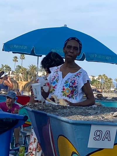 The image depicts a sunny day at a beach in Marbella, Spain. The primary subject is a person wearing a white shirt with colorful floral embroidery and a black headband. They are holding a white cup and appear to be interacting with someone or something off-camera. In the background, a blue umbrella with "UV Protection" written on it is visible. Further back, palm trees suggest a tropical or Mediterranean environment. There's also a sign with "9A" on it attached to a blue object. Other people are present in the scene, some seated and others partially visible, suggesting a gathering or a busy beach day. The overall atmosphere seems relaxed and casual, indicative of a leisure activity at the beach. The lighting suggests it is daytime.