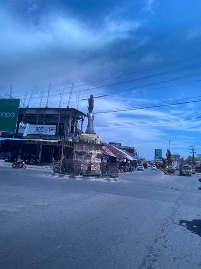 The image shows a street scene in Simpangpeut, Indonesia. In the foreground, a paved road curves to the right. In the middle ground, a roundabout features a monument with a sculpture of a figure topped with a corn cob. Surrounding the roundabout are several buildings, including a multi-story structure under construction with exposed rebar, and shops with corrugated metal roofs. A red scooter with two occupants is seen in the left part of the frame, navigating the roundabout. Further down the road, other vehicles including cars and a yellow truck are visible. Overhead, power lines crisscross the bright blue sky, which is partially obscured by clouds. A large green billboard with an image of a person is visible on the right side of the road in the distance. The scene depicts a busy urban environment with ongoing construction and daily traffic.