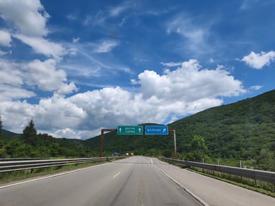 The image shows a highway scene with a bright blue sky filled with white, fluffy clouds. The road ahead splits, with two green and blue directional signs overhead. The left sign, green with white lettering and arrows, indicates "VARNA" straight ahead. The right sign, blue with white lettering and an arrow, points to "ZERKOVO". The highway is bordered by guardrails and lush green, tree-covered hills on both sides. In the distance, there are a few vehicles on the road. The overall impression is a scenic drive through a mountainous, forested region, likely on a highway leading to or from the indicated destinations. The location is stated as Potop, Bulgaria.