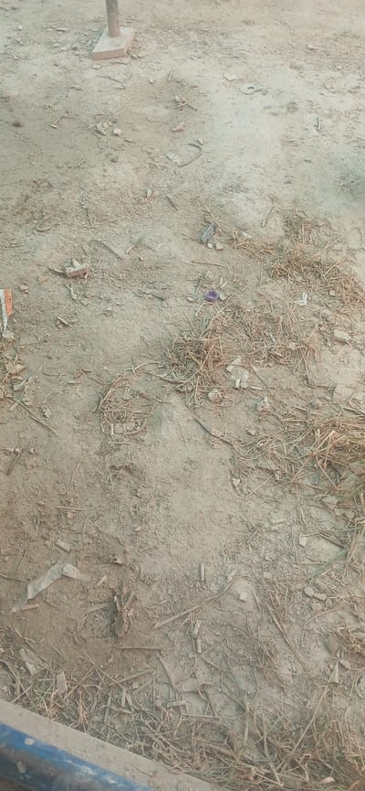 The image shows a close-up, overhead view of a dusty, outdoor ground surface littered with dry grass, small rocks, and debris. In the upper center, a thin metal pole is partially visible, resting on a light-colored rectangular base. Some pieces of broken concrete or brick are scattered around the base of the pole. There is also a small, purple plastic cap visible in the middle right portion of the image, nestled amongst the dry grass. The ground itself appears to be a mix of dirt and fine gravel. The lower part of the image shows a textured, possibly wooden or concrete structure with a blue element. The overall scene suggests an unkempt outdoor area, possibly in Daliguzar, Uzbekistan, with no people or animals present.