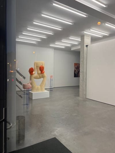 The image depicts an art gallery interior with a polished concrete floor. Several fluorescent light fixtures are mounted on the white ceiling. In the foreground, a large abstract sculpture, appearing to be made of a light-colored material with orange accents on its upper extremities and pink elements near its base, is displayed on a white plinth. Two smaller pink sculptures on blue metal stands are positioned to the left and right of the main sculpture. Further into the gallery, a concrete pillar is visible, and on the wall behind it, a framed artwork with warm tones is displayed. The scene is viewed through a glass storefront, with reflections of streetlights and a building facade visible on the glass. A metal trash bin is situated near the entrance. The location is identified as Saint-Gilles - Sint-Gillis, Belgium.