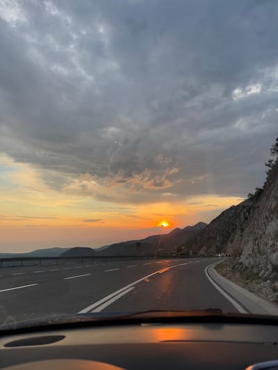 The image depicts a scenic highway passing through a mountainous coastal region, likely Brajici, Montenegro, during sunset. The road, with distinct white lane markings and a guardrail on the left, curves to the right. On the right side of the road, a steep, rocky mountainside with sparse vegetation rises, and in the distance, several vehicles are visible on the highway. The sky is filled with dramatic clouds, with the sun setting behind a mountain range on the left, casting a warm, orange glow that reflects on the wet road surface. The overall scene suggests a journey along a picturesque route at dusk.
