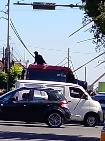 The image captures a street scene in Tabanan, Indonesia, under a clear blue sky. In the foreground, a black SUV is parked, and its driver is visible through the window. Behind it, a white van is also present. A red truck, with a person sitting on its roof, is further in the background. The scene is set against a backdrop of utility poles, power lines, and lush green trees. The overall impression is one of urban life on a bright, sunny day.