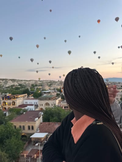 The image captures a scenic view of numerous hot air balloons ascending over a unique landscape, likely during sunrise or sunset, with a person observing the spectacle. In the foreground, the upper back and right side of a person are visible, featuring dark, tightly braided hair and a dark top. The person's head is slightly turned to the left, directing their gaze towards the panoramic view.

The sky is a clear gradient, transitioning from a soft blue at the top to a warm, pale orange and pink near the horizon, indicating either dawn or dusk. Dozens of colorful hot air balloons, some with distinct patterns, are scattered across the vast sky, appearing at various distances and altitudes.

Below the horizon, a sprawling town or village is visible, characterized by light-colored buildings with traditional architecture, nestled among rugged rock formations and green trees. The natural landscape of the region is evident, with some structures seemingly carved into the hillsides. This distinctive setting, combined with the abundance of hot air balloons, strongly suggests the location is Göreme, Turkey (Cappadocia). The overall atmosphere is serene and awe-inspiring. No readable text is visible in the image.