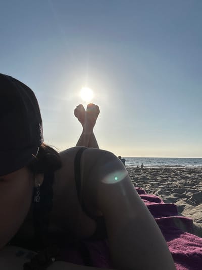 The image displays a female individual lying prone on a sandy beach in Manhattan Beach, United States. The subject is partially visible, primarily her left side, dressed in a dark swim top and a dark baseball cap that obscures her face. Her feet are elevated and crossed at the ankles, silhouetted against a clear, light blue sky, with the bright, low-lying sun positioned directly between them. The sun casts significant backlighting, creating shadows on the foreground subject and exhibiting a green-blue lens flare. The individual rests on a purple towel, partially visible beneath her. The Pacific Ocean forms the background, featuring a distinct horizon line and gentle waves. A faint, dark silhouette of another person is discernible in the distant background near the water's edge.