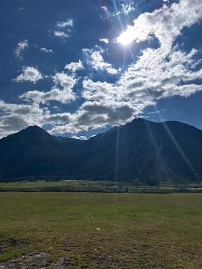 A wide-angle, vertical photograph depicts a mountain range under a bright, partly cloudy sky. The sun is visible in the upper right quadrant of the sky, casting rays of light through the clouds. The mountains, covered in dark green trees, dominate the background. In the foreground, a vast, grassy field stretches out, with a dark patch of earth in the lower left corner. A wooden fence runs horizontally across the middle ground, separating the field from a distant treeline and a few scattered buildings. The overall impression is one of a serene, natural landscape.