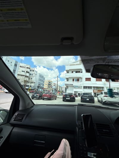 Captured from within a vehicle, the image shows an urban scene in Famagusta, Northern Cyprus/Cyprus. Through the windshield, buildings are visible, including white residential structures and taller, multi-story buildings. Parked cars line the street. In the foreground, the car's dashboard and rearview mirror are prominent. A phone is mounted on the dashboard. The sun visor is partially lowered. The sky is blue with scattered clouds.