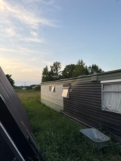 In the foreground, a dark, corrugated surface leans diagonally from the left edge of the frame, appearing to be part of a structure, possibly solar panels. Behind this, tall, green grass covers the ground. A mobile home, with a two-tone exterior of light green and dark grey horizontal siding, is situated in the middle ground. The mobile home has several windows, two of which are partially open, revealing white curtains in one. To the right of the mobile home and in the grass, a metal wheelbarrow is visible. In the background, a field stretches towards a line of trees and a few houses. The sky above is a pale blue with wispy white clouds, indicating either early morning or late evening. The scene appears to be in a rural or semi-rural setting, possibly on the outskirts of Vol'noe, Russia. There are no people or animals visible in the image.