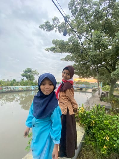 Two young girls are standing outdoors on the bank of a waterway in Kalitengah, Indonesia. The girl in the foreground on the left wears a light blue dress with a dark blue hijab, facing forward with a slight smile and her mouth slightly open. Her hands are visible at her sides. The girl behind her on the right wears a tan long-sleeved top with a brown skirt and a brown hijab, accented with a red and white scarf. She is looking towards the camera with a subtle smile.

Behind the girls, a calm body of water, likely a river or canal, reflects the overcast sky. On the opposite bank, light-colored walls with colorful murals are visible, alongside scattered green trees. Further in the background, a light blue bridge spans the waterway, and the sky shows an orange glow near the horizon, indicative of a sunrise or sunset. To the right of the girls, dense green foliage and a tree with green leaves are present. String lights are strung overhead between the tree and an unseen anchor point. The ground beneath the girls appears to be a concrete or paved pathway.
