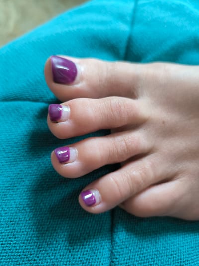 The image displays a close-up view of a person's toes, which have been painted with a dark purple nail polish. The toes are resting on a teal-colored textured fabric, possibly a blanket or upholstery. The lighting suggests an indoor environment. There is no discernible information within the image to indicate a specific location in Miass, Russia. The focus is solely on the painted toenails and the fabric they are on.