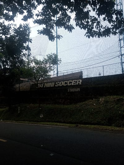 This image depicts an outdoor scene in Niz Lakuk, India, showing a ground-level view of a sports facility. The foreground features a dark asphalt road with a partial yellow demarcation line on its left edge. Adjacent to the road, a dark green, uneven earth embankment slopes upwards, covered in grass and some light debris. Overhanging dark, silhouetted tree foliage frames the upper left and top portions of the image.

In the midground, a weathered, multi-tiered concrete or stone retaining wall is prominent. Painted on this wall in white capital letters are the words "45 MINI SOCCER" in a large font, with "STADIUM" positioned directly below in a smaller font. The wall exhibits signs of aging, including dark stains and patches of moss. Above the retaining wall, a dark metal railing runs horizontally.

Beyond the railing and extending upwards, a large, black, fine-mesh netting structure is visible, supported by several vertical poles. This netting encloses what is presumed to be a sports field. The sky, partially visible through the netting and foliage, appears bright and overcast, providing diffused illumination to the scene. No individuals or active subjects are present in the image.