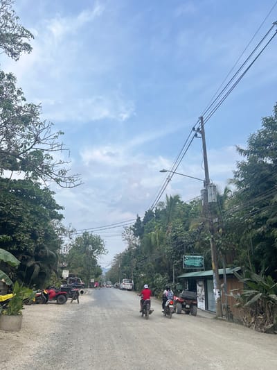 The image shows a street in Santa Fe, Costa Rica, with a clear blue sky above. The street is lined with lush green trees and vegetation on both sides. Multiple power lines with a utility pole in the center stretch across the road. There are numerous parked vehicles, including a red four-wheeler with two people sitting on it, and two motorcycles, as well as a van and several cars. Two people are riding motorcycles down the road. A sign for a business, "D'CENA" is visible on the right side of the image. The road is dusty and the vegetation is dense, with large trees and bushes. The image appears to have been taken on a bright, sunny day.