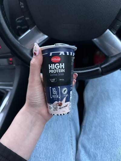 In Rathvilly, Ireland, a person's hand, adorned with intricate nail art featuring flowers and ladybugs, holds a Milbona High Protein Coffee Drink. The hand is positioned near a car's steering wheel, suggesting the photo was taken from inside a vehicle. The coffee drink is in a black and blue container with text indicating "19g PROTEIN per cup," "No added sugars," and "Low fat." The background shows parts of a car interior, including the steering wheel and dashboard, with hints of a person's legs dressed in denim. The lighting suggests an indoor setting, possibly a car parked in daylight.