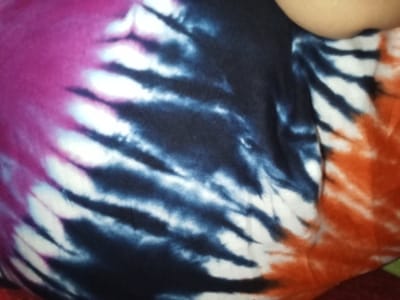 The image displays a close-up, oblique view of a multi-colored tie-dye fabric, likely clothing, situated in Ciputri, Indonesia. The fabric features a radial or spiral pattern incorporating sections of magenta, dark indigo, white, and orange/red. A partial, light-toned human upper arm or shoulder is visible in the upper right quadrant, indicating the fabric is either worn by or in direct contact with an individual. The fabric exhibits minor creases and folds.