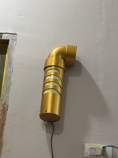 A gold-colored outdoor light fixture is mounted on a grey wall. The fixture is cylindrical with horizontal bands and a bent pipe-like top. A black electrical cord extends from the bottom of the fixture. To the left of the light, a portion of a doorway with peeling paint is visible. To the lower right of the light, a light switch with two toggles is mounted on the wall. The scene appears to be an exterior wall in Tupah, Indonesia.