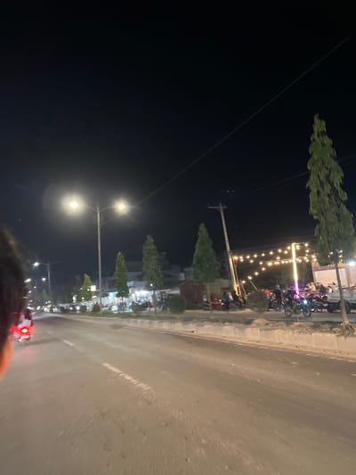 This media file depicts a nighttime scene on a street in Bundar, Indonesia. The perspective is from someone riding a motorcycle, as indicated by the red taillight visible in the foreground. The street is paved and appears wet, suggesting recent rain or ongoing damp conditions.
Along the side of the road, there are several parked motorcycles, indicating a gathering or a place of interest. A line of decorative string lights illuminates the scene, creating a festive and inviting atmosphere. Several trees, some conical in shape and others more mature and leafy, are planted along the roadside, adding greenery to the urban landscape. Streetlights are also present, casting bright pools of light.
In the background, buildings are visible, suggesting a commercial or residential area. People are present, some on motorcycles and others near the buildings, likely socializing or engaging in activities related to the establishments. The overall mood seems lively and social, despite the darkness of the night. The presence of motorcycles and the roadside setting are common in Indonesia. The time of day is clearly night, indicated by the darkness and artificial lighting.