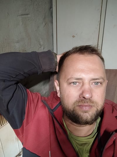 A man with short brown hair and a beard, wearing a red and black zip-up hoodie over a green t-shirt, is looking directly at the camera. His left arm is raised, with his hand resting on the back of his head. He is indoors, in what appears to be a room with a textured, light-colored wall to his left and a lighter, vertical panel to his right. The background suggests an interior setting, possibly a residence or a utility space, with no discernible identifying features that would pinpoint a specific location within Salzgitter, Germany. The lighting is somewhat dim, creating shadows on the wall.