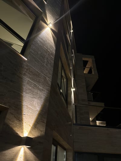 The image shows the exterior of a modern building at night in Tyulenovo, Bulgaria. The building features a facade clad in light-colored, horizontally striated stone or tile. Several exterior lights are mounted on the wall, casting dramatic upward and downward beams of warm light. These lights illuminate portions of the stone, creating contrasting areas of light and shadow. In the background, the dark night sky is visible, with no stars or moon apparent. The architecture includes geometric shapes and what appears to be a balcony or open-air section with a glass railing. One section of the building features an angular, abstract architectural element that is also illuminated by the lighting. The overall impression is one of modern, minimalist design emphasized by dramatic nighttime illumination. There are no people or animals visible in the image.