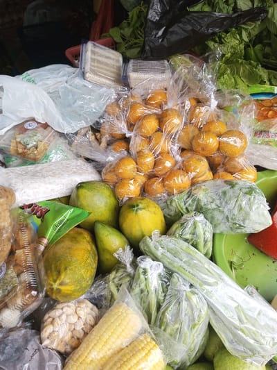 The scene depicts a tightly packed arrangement of assorted produce and packaged goods, likely on a market stall surface, located in the city of Bagochen Boolingpang Ueling, Bhutan. The upper portion of the image displays dark background elements, including a black plastic bag and a cluster of green leafy vegetables. Towards the upper center, several clear plastic containers hold light-colored, rectangular food items. A prominent central feature is a large collection of small, round, orange citrus fruits, each individually contained within a clear plastic bag, with these smaller bags then clustered together. To the mid-left, a white plastic bag holds numerous light-colored grains or beans, adjacent to a clear plastic container with fried snacks and a partially visible green snack bag. In the lower-middle and mid-right sections, several papayas, varying from green to yellow-green in ripeness, are present alongside numerous bundles of various green vegetables, including leafy greens and long slender varieties, all encased in clear plastic packaging. A bright green plastic basin and a section of a red object are partially visible on the far right. The bottom left features a clear plastic bag containing nuts, and at the bottom center, two ears of husked yellow corn are displayed. No individuals are visible within the frame.