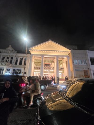 The image captures a nighttime scene in Medan, Indonesia, focusing on a brightly lit department store with a classical facade. The store, identified by signs reading "SENG HAP 1881" and "SAWAN," features grand columns and illuminated architectural details, suggesting a grand opening or a busy shopping evening.
Several people are visible, both inside and outside the store, with some browsing merchandise and others interacting on the sidewalk. A black car is parked in the foreground, partially obscuring the view of the building and reflecting the store's lights. Another vehicle, possibly a motorbike, is also present, with a blurry figure on it. The overall atmosphere appears lively, with the warm lighting of the store contrasting with the dark night sky. A street lamp shines brightly in the upper left quadrant of the image, adding to the nighttime illumination. The architecture of the building, with its prominent columns and pediment, gives it a sense of historical significance. The presence of people and vehicles suggests an active urban environment.