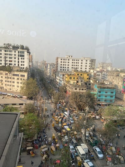 The image presents an elevated, wide-angle view of a densely populated urban street in Narayanganj, Bangladesh, under hazy daylight conditions. The vantage point appears to be from a building, with reversed text "Shoe w" and window frame reflections visible in the upper portion of the frame, indicating the photograph was taken through glass.

A long, straight asphalt road bisects the image, extending from the foreground into a light blue, hazy distance. Both sides of the road are lined with multi-story buildings, typically ranging from three to seven stories in height, featuring varied architectural styles and color schemes, including white, beige, yellow, and light blue. Several rooftops exhibit vegetation or clutter.

The road itself is heavily congested with traffic. Dominant vehicles include numerous yellow three-wheeled auto-rickshaws (CNGs), along with cars (such as a white van and a silver sedan), and smaller trucks/vans. Pedestrians are abundant on sidewalks and are observed crossing the street. A dense network of overhead utility wires crisscrosses the scene, supported by numerous utility poles along the road. Sparse trees with minimal foliage are interspersed between buildings and along the street. The overall environment depicts a bustling, crowded urban landscape.